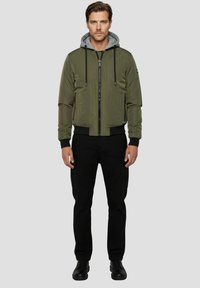 Grüne Bomberjacke mit grauer Kapuze, Reißverschluss, seitlichen Taschen und gerippten Bündchen; kombiniert mit schwarzen Hosen und schwarzen Schuhen.