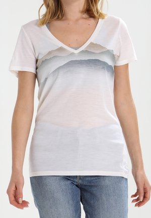 Frau trägt weißes V-Ausschnitt-T-Shirt mit grauem und beigem Bergmotiv, kombiniert mit hellblauen Jeans, stehend vor weißem Hintergrund.