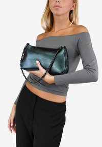 Bolso de mano de cuero negro con una correa de cadena, con un acabado suave y diseño minimalista. Sostenido en una mano contra un fondo liso.