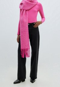Foulard long fuchsia à franges, porté sur un haut à manches longues rose côtelé. Jean noir à jambes larges et bottines noires pointues à talons complètent la tenue.