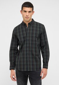 Mann trägt ein dunkelgrünes und schwarzes karierter Button-Up-Hemd mit langen Ärmeln und steht vor einem schlichten hellen Hintergrund.