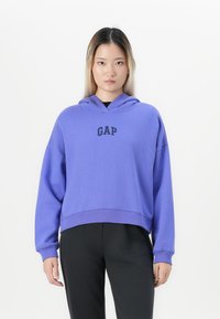Jeune personne portant un sweat à capuche violet de GAP et un pantalon noir, se tenant devant un fond blanc uni, faisant face à l'avant.
