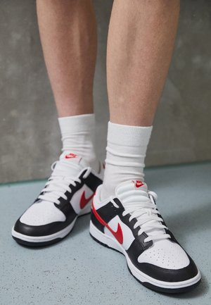 Jambes portant des chaussettes blanches et des baskets Nike noires, blanches et rouges, debout sur un sol bleu clair tacheté avec un mur gris en arrière-plan.