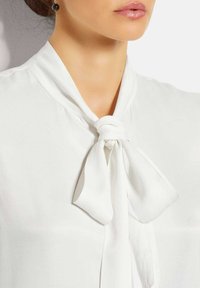 Weißes Blusenshirt mit einem Bandkragen, aus glattem Stoff und weicher Textur. Das Band ist in der Mitte gebunden und schafft einen fließenden Fall.