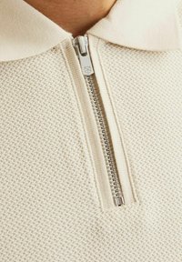 Svetlo béžová polo košeľa s textúrovaným vzorom, ktorá má krátky zips a pletený golier. Zips má strieborný kovový pull.