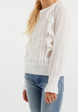 Frau trägt eine weiße, strukturierte Bluse mit Rüschen und gerafften Manschetten, kombiniert mit hellblauen Jeans, steht vor einfachem Hintergrund.