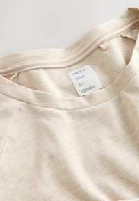 Helles beiges T-Shirt mit weicher Textur, mit Rundhalsausschnitt und einem Etikett, das "NÄCHSTE Mal zum Träumen" liest. Keine auffälligen Muster oder Akzente.