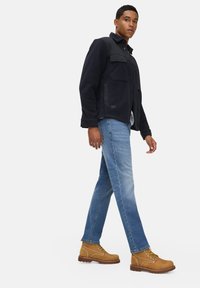 Donker marineblauwe jas met twee borstzakken en een gladde textuur, gecombineerd met lichtblauwe jeans en tan kleurige veterschoenen.