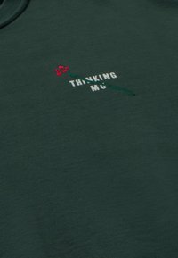 Tmavo zelená tkanina s bielym vyšívaným textom "THINKING MU", prekrížená malou vyšívanou červenou kvetinou so zeleným stonkom.