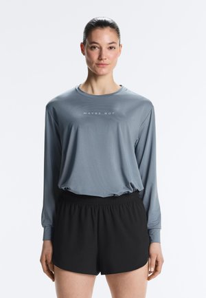 Ženska v sivi majici z dolgimi rokavi z napisom "MAYBE NOT" in črnih kratkih hlačah z elastičnim pasom, stoji pred enobarvnim belim ozadjem.