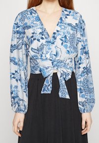 Blusa con motivo floreale blu e bianco, scollatura a V profonda, maniche lunghe e un dettaglio in vita annodato, abbinata a una gonna nera.