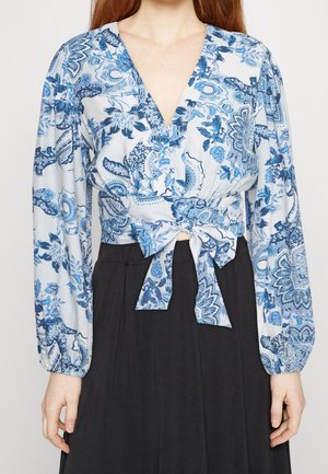 Blouse à motif floral bleu et blanc avec un décolleté en V plongeant, des manches longues et un détail de taille cintrée, associée à une jupe noire.