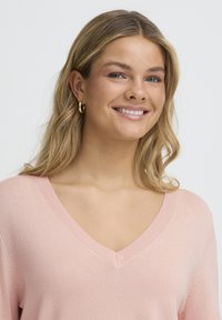 Pull rose clair à col en V avec encolure côtelée et manches courtes. Texture douce. Le modèle a des cheveux ondulés et porte de petites boucles d'oreilles créoles en or.