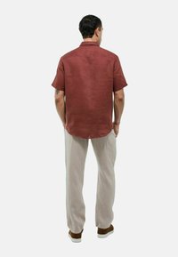 Chemise à manches courtes pour homme en lin de couleur rouille, vue de dos. Associée à un pantalon beige clair et des baskets marron. Texture lisse, coupe décontractée.