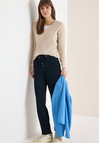 Femme aux cheveux bruns portant un pull beige et un pantalon bleu marine, tenant une veste texturée bleu vif, debout devant un fond blanc et beige.