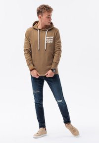 Felpa color khaki con lettering bianco sul petto, cordini, tasca a marsupio; abbinata a jeans scuri strappati e sneakers beige.
