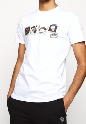 Biała koszulka z pięcioma nadrukami w stylu taśmy na klatce piersiowej, noszona przez osobę z rękami w kieszeniach czarnych spodni.