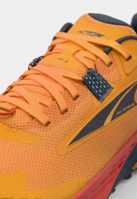 Chaussure de sport orange avec un dessus en maille texturée, des accents sombres contrastants, des lacets fins et une semelle à motif. Présente un logo visible.