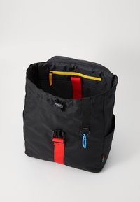 Schwarzer Rucksack mit einem strukturierten Stoffaußenmaterial, ausgestattet mit einem roten Riemen, einer gelben Innenreißverschlusstasche und blauen Akzenten. Kompakte quadratische Form.