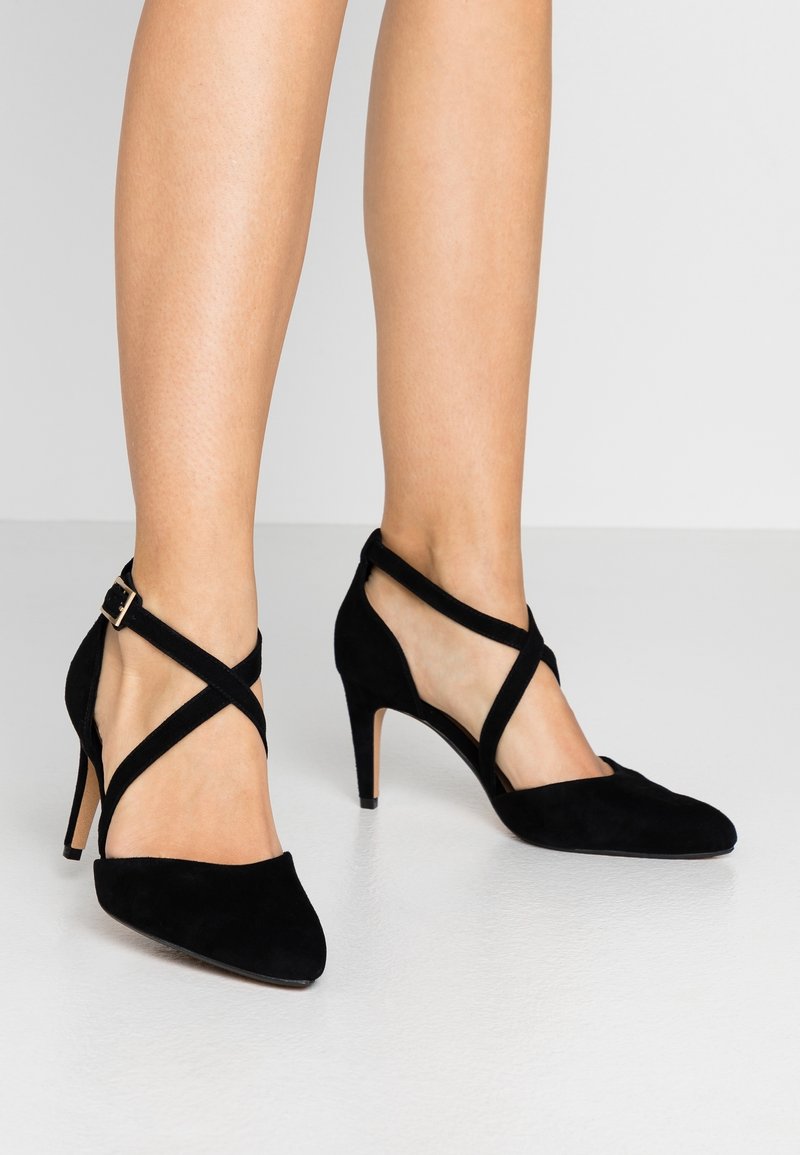 Jambes féminines portant des chaussures à talons hauts en daim noir avec des brides croisées à la cheville et des bouts pointus sur un sol lisse et clair.