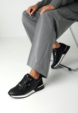 Černé tenisky s texturovaným svrškem a zlatými detaily, spárované se šedými pouzdrovými kalhotami. Mají bílou podrážku a prošití.