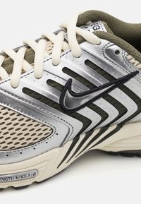 Nike löparskor med ett silver- och olivgrönt nätöverdelse, svarta accenter, krämfärgade snören och en texturerad sula med synlig märkning.