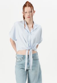 Camisa cropped listrada azul clara com mangas curtas, botão na frente, detalhe de nó na cintura, combinada com jeans de denim claro de cintura alta.