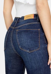 Donkerblauwe spijkerbroek van denim met contrasterende oranje stiksels, voorzien van twee achterzakken en een leren merketiket op de tailleband.