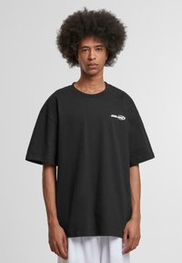 Black oversized cotton t-shirt with a round neck and short sleeves. Features a white logo on the left chest area. Smooth texture.