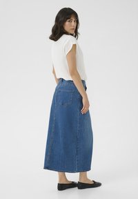 Jupe maxi en denim bleu avec une texture lisse, taille haute et poches arrière. Associée à un blouse blanche à manches courtes.