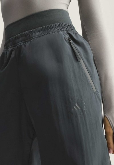 Nahaufnahme einer Person, die dunkelgraue, elastische Bundhose trägt, mit einer Reißverschlusstasche und einem schwach sichtbaren runden Logo in der Nähe des Oberschenkels.