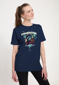 Žena s dlouhými vlasy v culíku, která má na sobě námořnickě modré tričko s barevnými sluchátky a textem ve stylu graffiti "Straightfire Tropical Twist."