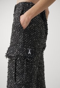 Zwart-witte tweed broek met een gestructureerde afwerking, voorzien van zijzakken en een zichtbaar merketiket aan de onderrand van de zak.