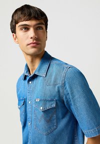 Verbleekte blauwe spijkerblouse met korte mouwen, borstzakken en contrasterende stiksels; klassiek ontwerp met yoke in westernstijl en knoopsluiting.