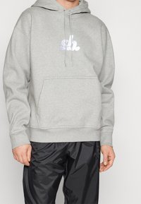 Hombre vistiendo una sudadera ligera gris con un logotipo bordado en blanco y pantalones de chándal negros brillantes, de pie contra un fondo neutro.