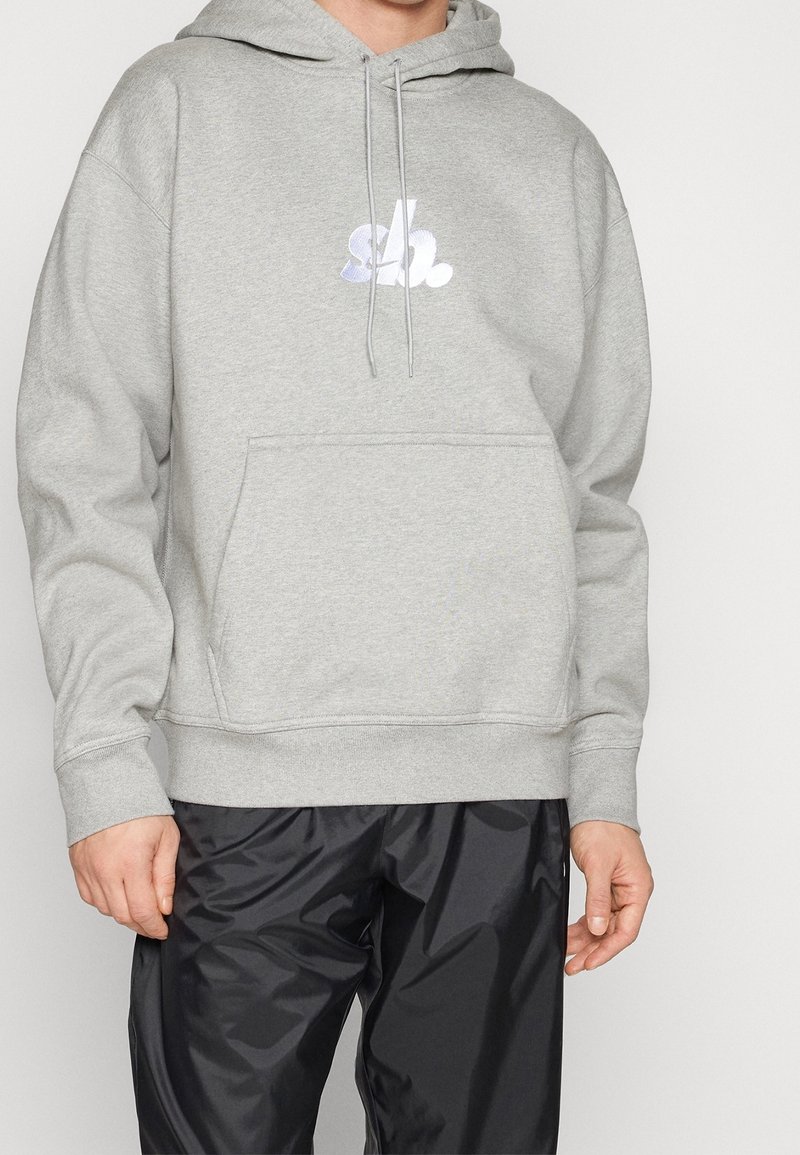 Hombre vistiendo una sudadera ligera gris con un logotipo bordado en blanco y pantalones de chándal negros brillantes, de pie contra un fondo neutro.