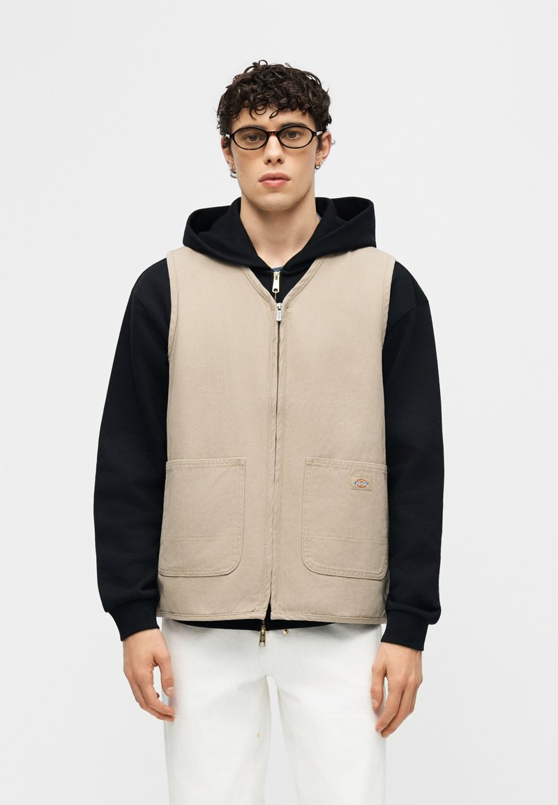 Joven con cabello rizado y gafas, lleva un chaleco beige con cremallera sobre una sudadera negra y pantalones blancos, de pie frente a un fondo liso.