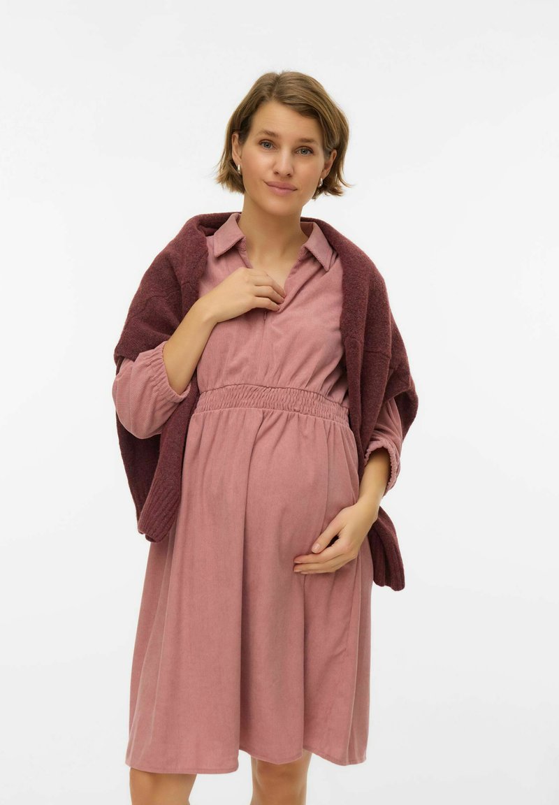 Porodniška obleka iz mehke roza tkanine s poudarjenim pasom, dolgimi rokavi in ovratnim izrezom, obložena z bordo pletenim šalom.