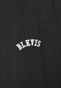 Țesătură neagră cu text brodat alb care citește "BLKVIS" într-un design curbat. Textura pare netedă cu o strălucire subtilă.