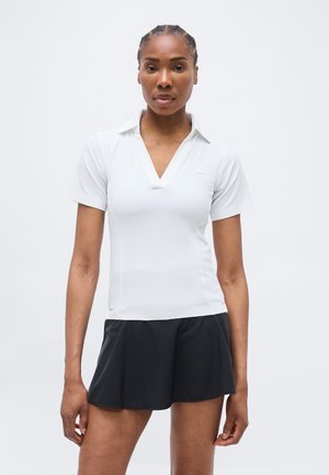 Nő áll, előre néz, fehér rövid ujjú, V-nyakú pólót és fekete sportnadrágot visel, egy egyszerű háttér előtt.