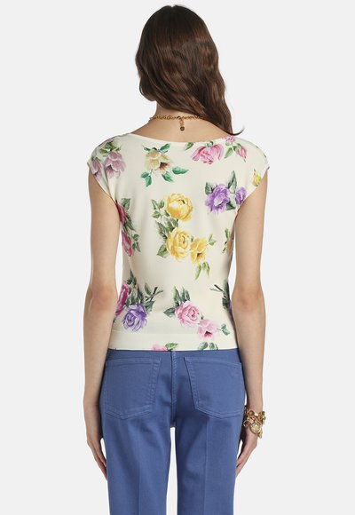 Frau mit langen braunen Haaren, die ein weißes, kurzärmeliges, florales Oberteil und blaue Hosen trägt, von hinten gezeigt, geschmückt mit goldenen Schmuckstücken.