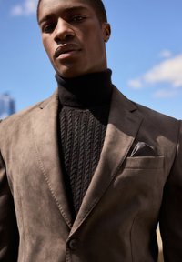 Blazer in suede marrone sovrapposto a un dolcevita nero a coste, con un fazzoletto da taschino e dettagli di cucitura discreti. Sfondo con cielo blu chiaro.