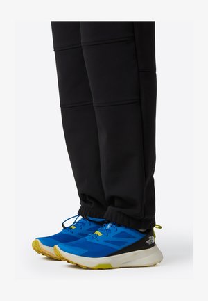 Blauwe sportschoenen met een bovenwerk van mesh, gele accenten en een witte zool, gecombineerd met zwarte, nauwsluitende trainingsbroeken met elastische boorden.