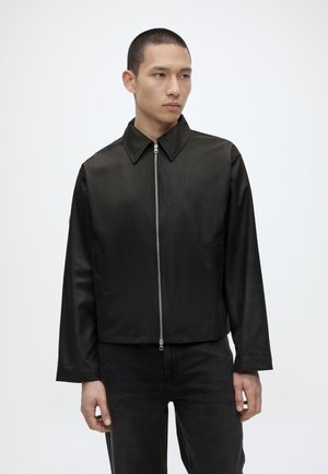 Man met kort haar, gekleed in een zwarte jas met rits en een zwarte broek, staand tegen een effen lichte achtergrond.