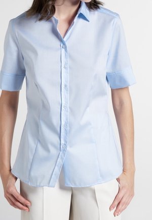 Vrouw die een lichtblauw kortemouwen overhemd met knopen en een witte broek draagt, staat met haar handen ontspannen langs haar zij.