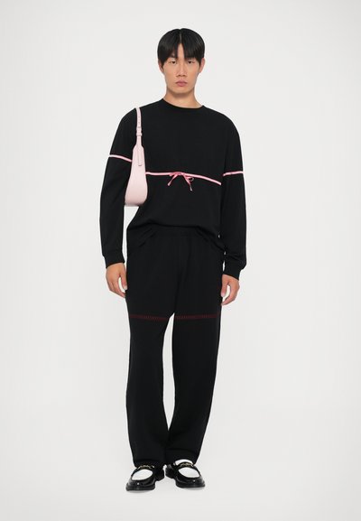 Maglietta nera a maniche lunghe con dettagli rosa e un fiocco, abbinata a pantaloni neri con cuciture rosse. Il modello tiene in mano una piccola borsa rosa.