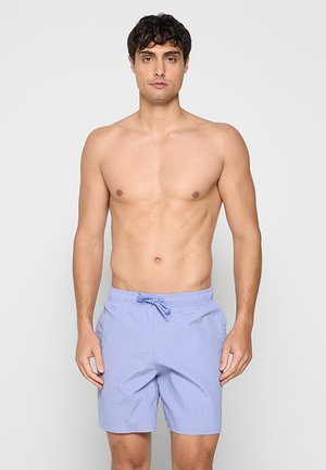 Oberkörperfreier Mann mit kurzen lockigen Haaren, der hellblaue Badehosen mit Kordelzug trägt und vor einem einfarbigen hellgrauen Hintergrund steht.