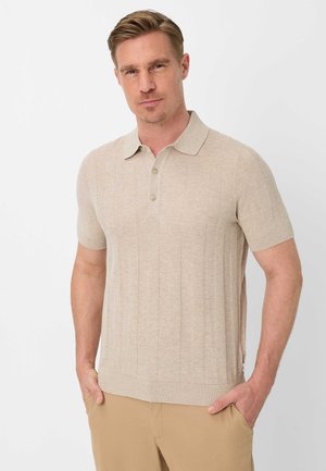 Mand med kort blondt hår iført beige kortærmet strikket polo og kaki bukser, hænderne i lommerne, neutral baggrund.