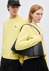 Άνδρας και γυναίκα φορούν ταιριαστά κίτρινα πουλόβερ και μαύρο παντελόνι· η γυναίκα κρατά μια μεγάλη μαύρη τσάντα με πιέτες στον ώμο της.