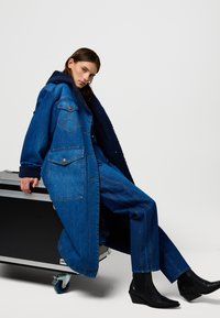 Płaszcz dżinsowy z puszystym granatowym kołnierzem, z naszywanymi kieszeniami i zapięciem na guziki. Modelka ma na sobie ciemne, zwężane spodnie oraz czarne botki na koturnie.